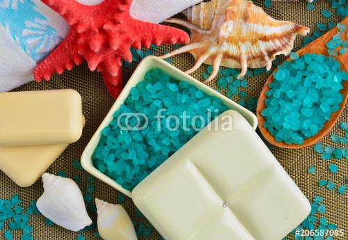 Blue sea salt for spa treatments in a white square ceramic box with a lid, next to white terry towels, a red starfish and seashells