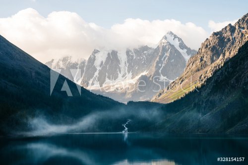Beautiful big glacier, rocky snowy mountains, coniferous forest on hills, mountain lake and highland creek under cloudy sky. Atmospheric alpine scenery with low clouds on high forest steep