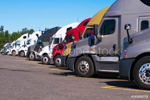 Big rigs semi trucks of different makes and models stand in row on truck stop parking lot