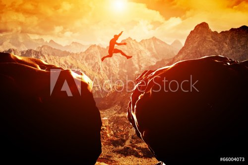 Man jumping over precipice between two mountains at sunset