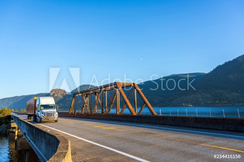 White big rig semi truck transporting cargo in orange semi trailer driving on the bridge across the Columbia River