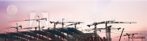 The silhouette image of the metal truss steel structure of the building construction project with the operation of a group of tower cranes and the background of golden sky of sun set.