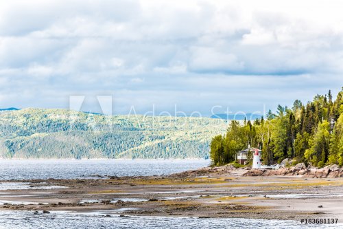 Fjord coast nature near Saguenay river, cliffs, tree forest, mountains and cloudy clouds sky, sunset light and small red lighthouse