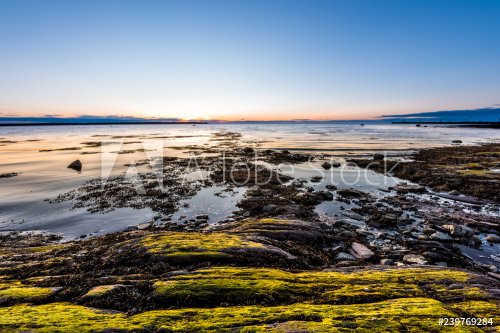 Sunset, dusk in Rimouski, Quebec, Saint Lawrence river, Gaspesie, Canada with rocks, boulders, rocky beach, turquoise water, sun reflection above horizon, sunburst, glade, path, seaweed blue sky