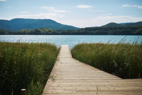Long wooden pier leads through tall grass to a sparkling blue lake surrounded by wooded hills on a clear summer day
