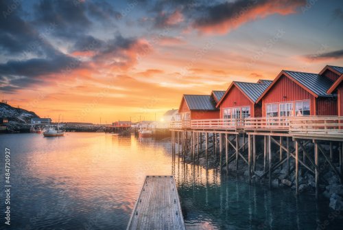 Red rorbu on wooden piles on sea coast, small jetty, colorful orange sky at sunrise in winter. Lofoten islands, Norway.
