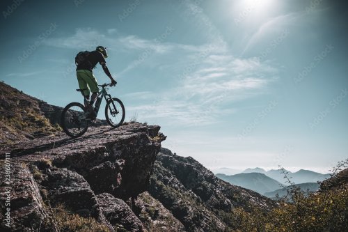 Mountain biker riding up a large rock deep in the alps with sun and mountain layers behind